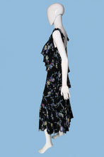 Load image into Gallery viewer, A black sleeveless dress with a colorful floral print, displayed on a white mannequin. The dress features a V-neckline, ruffled shoulder straps, and a tiered ruffle detail at the waist.
