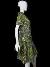 Load image into Gallery viewer, A short-sleeve collared dress with a vibrant green, yellow, purple, and blue paisley pattern, displayed on a white mannequin against a plain wall.
