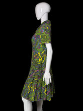 Load image into Gallery viewer, A short-sleeve collared dress with a vibrant green, yellow, purple, and blue paisley pattern, displayed on a white mannequin against a plain wall.
