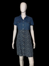 Load image into Gallery viewer, A short-sleeve button-front dress displayed on a white mannequin. The top portion is medium blue denim, and the skirt portion has a small repeating floral or paisley-like pattern in blue, white, and dark tones.
