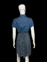Load image into Gallery viewer, A short-sleeve dress displayed on a white mannequin, viewed from the back. The upper portion is solid medium blue denim-like fabric, while the skirt portion has a small-scale blue and white floral.
