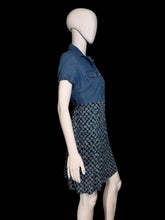 Load image into Gallery viewer, A denim shirt dress displayed on a white mannequin against a plain indoor wall. The top portion is solid medium blue denim, while the skirt portion has a small-scale floral print in dark blue and white.
