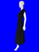 Load image into Gallery viewer, A black midi-length dress displayed on a white mannequin in a room corner. The dress has short sleeves with gold chain detailing along the shoulders and a front zipper.
