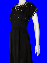 Load image into Gallery viewer, A black short-sleeve dress displayed on a white mannequin against a plain light background. The dress features gold-tone coin embellishments along the neckline and sleeve cuffs, with additional gold coins scattered on the bodice.
