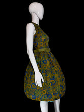 Load image into Gallery viewer, A sleeveless knee-length dress with a pleated skirt and matching belt, featuring a bold floral pattern in olive green, mustard yellow, and blue, displayed on a white mannequin.
