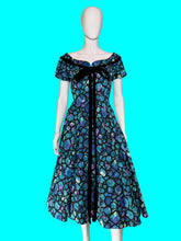Load image into Gallery viewer, A short-sleeve off-the-shoulder dress displayed on a white mannequin, featuring a vibrant blue, purple, and green abstract pattern with a black center panel and black bow detail.
