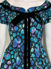 Load image into Gallery viewer, A 1950s off-the-shoulder dress displayed on a white mannequin. The dress features a vibrant abstract organic pattern in shades of blue, purple, and teal outlined in black, with a prominent black velvet bow at the neckline.
