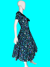 Load image into Gallery viewer, A vintage 1950s-style A-line dress displayed on a white mannequin. The dress features a vibrant blue, purple, and green abstract organic pattern with black trim and a black bow at the neckline.

