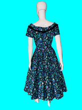 Load image into Gallery viewer, A vintage 1950s-style A-line dress displayed on a white mannequin. The dress features a vibrant blue, purple, and green abstract organic pattern with black trim and a black bow at the neckline.
