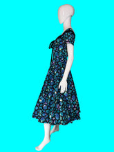 Load image into Gallery viewer, A vintage 1950s-style A-line dress displayed on a white mannequin. The dress features a vibrant blue, purple, and green abstract organic pattern with black trim and a black bow at the neckline.
