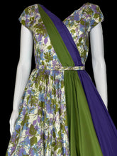 Load image into Gallery viewer, A short sleeve wrap-style dress displayed on a white mannequin, featuring a floral print bodice and skirt in cream, green, and purple, with a solid green and purple draped sash detail across the front.
