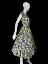 Load image into Gallery viewer, A short sleeve wrap-style dress displayed on a white mannequin, featuring a floral print bodice and skirt in cream, green, and purple, with a solid green and purple draped sash detail across the front.
