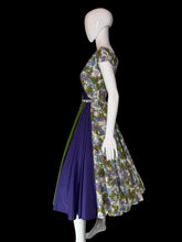 Load image into Gallery viewer, A short sleeve wrap-style dress displayed on a white mannequin, featuring a floral print bodice and skirt in cream, green, and purple, with a solid green and purple draped sash detail across the front.
