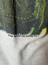 Load image into Gallery viewer, Close-up of a vintage Captain Kidd Clothes t-shirt showing the lower portion of a green and black graphic print with a white hem and copyright text reading '© 1989 CAPTAIN KIDD CLOTHES INC.'.
