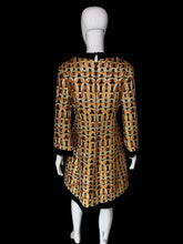 Load image into Gallery viewer, A women's tunic dress shown on a white mannequin. The dress features a bold geometric chain-link pattern in orange, black, and beige, with solid black trim at the split neckline, cuffs, and hem.

