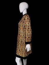 Load image into Gallery viewer, A women's tunic dress shown on a white mannequin. The dress features a bold geometric chain-link pattern in orange, black, and beige, with solid black trim at the split neckline, cuffs, and hem.
