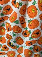 Load image into Gallery viewer, A close-up of a white fabric with a repeating pattern of orange pumpkins in various carved and smiling faces, accented with green leaves and vines.
