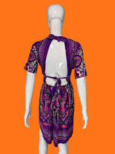 Load image into Gallery viewer, A back view of a purple and pink paisley print short-sleeve dress displayed on a white mannequin, featuring a high neck, tie details at the shoulders and waist, and a back cutout.
