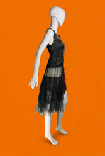 Load image into Gallery viewer, A 1920s drop waist sleeveless black dress with sheer ruffled layered skirt shown on a white mannequin from the side. 
