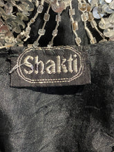 Load image into Gallery viewer, Close-up of a black fabric garment with a sewn-on brand label reading 'shakti' in white embroidery, next to clear beaded strands.
